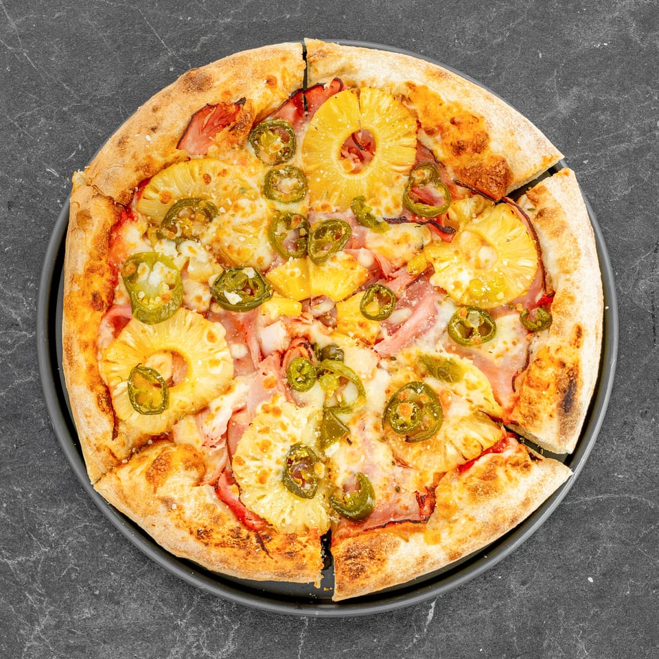 Overhead view of a sliced Hawaiian pizza topped with ham, pineapple, jalapeños, tomato sauce, and melted cheese on a black plate.