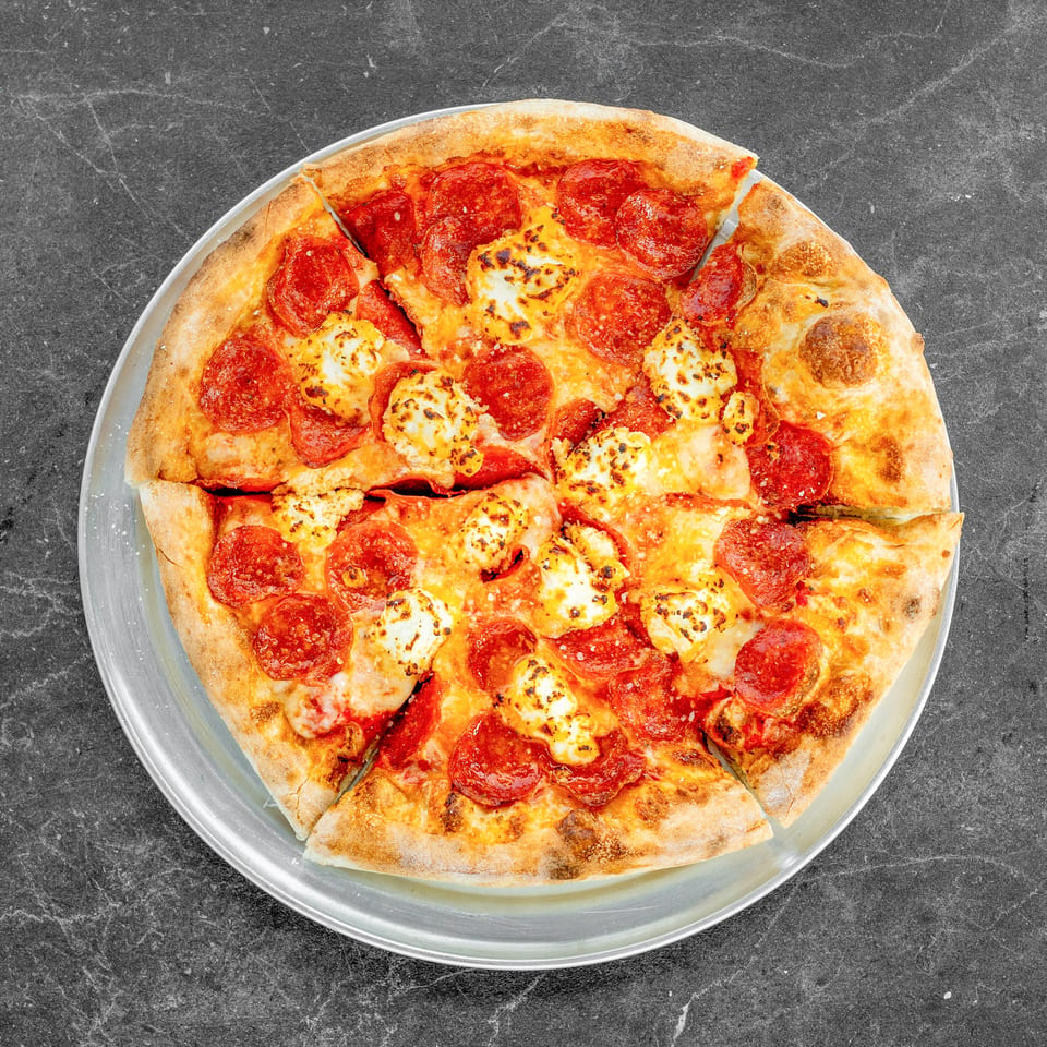 Overhead view of sliced pepperoni pizza with browned cheese on a round metal tray atop a dark stone surface.