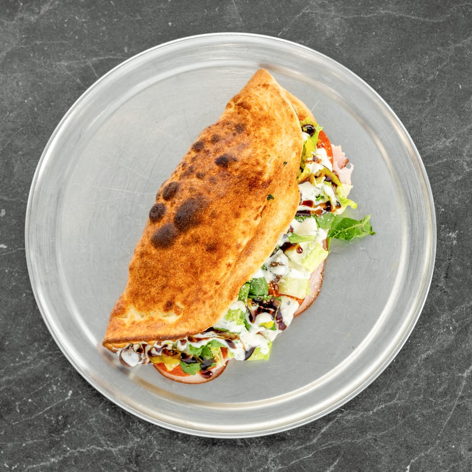 Golden-brown folded flatbread filled with leafy greens, vegetables, sliced meat, and creamy sauces on a metal plate.