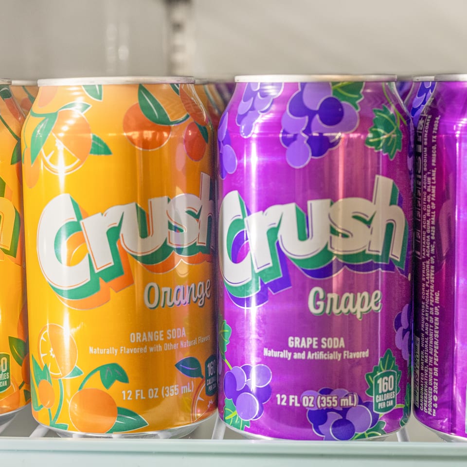 Rows of brightly colored orange and grape soda cans with fruit illustrations arranged on a refrigerator shelf.